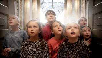 Group of children in different clothes standing together, indoors with gold columns in the background.