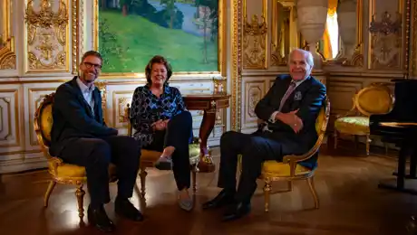 Two men and a woman are sitting on chairs in a richly decorated room.