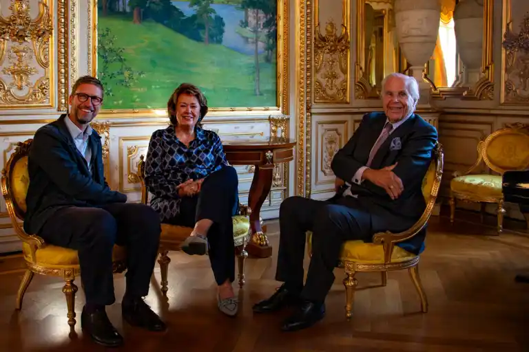 Two men and a woman are sitting on chairs in a richly decorated room.