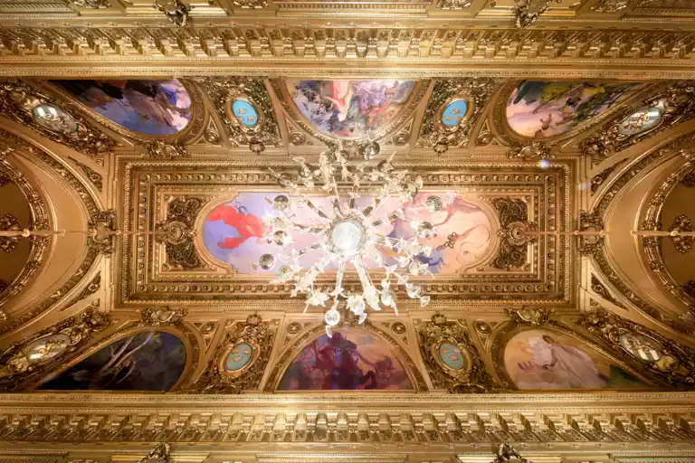 View of a richly decorated ceiling with gold and paintings, in the center a large chandelier.
