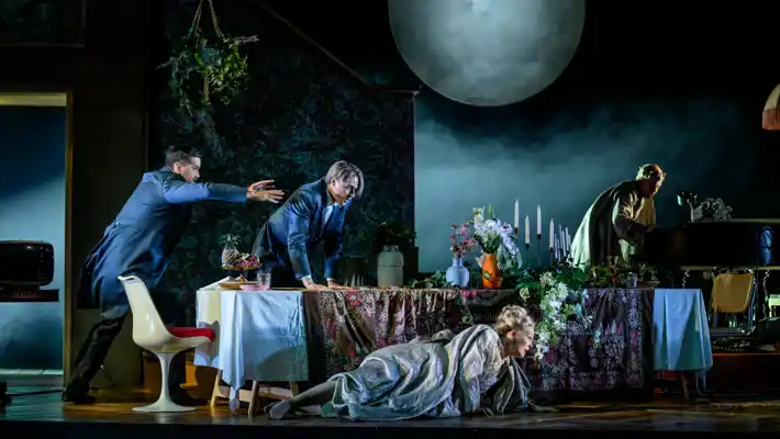 A scene with a set table, a woman lies on the floor in front of the table, several men move in the background.