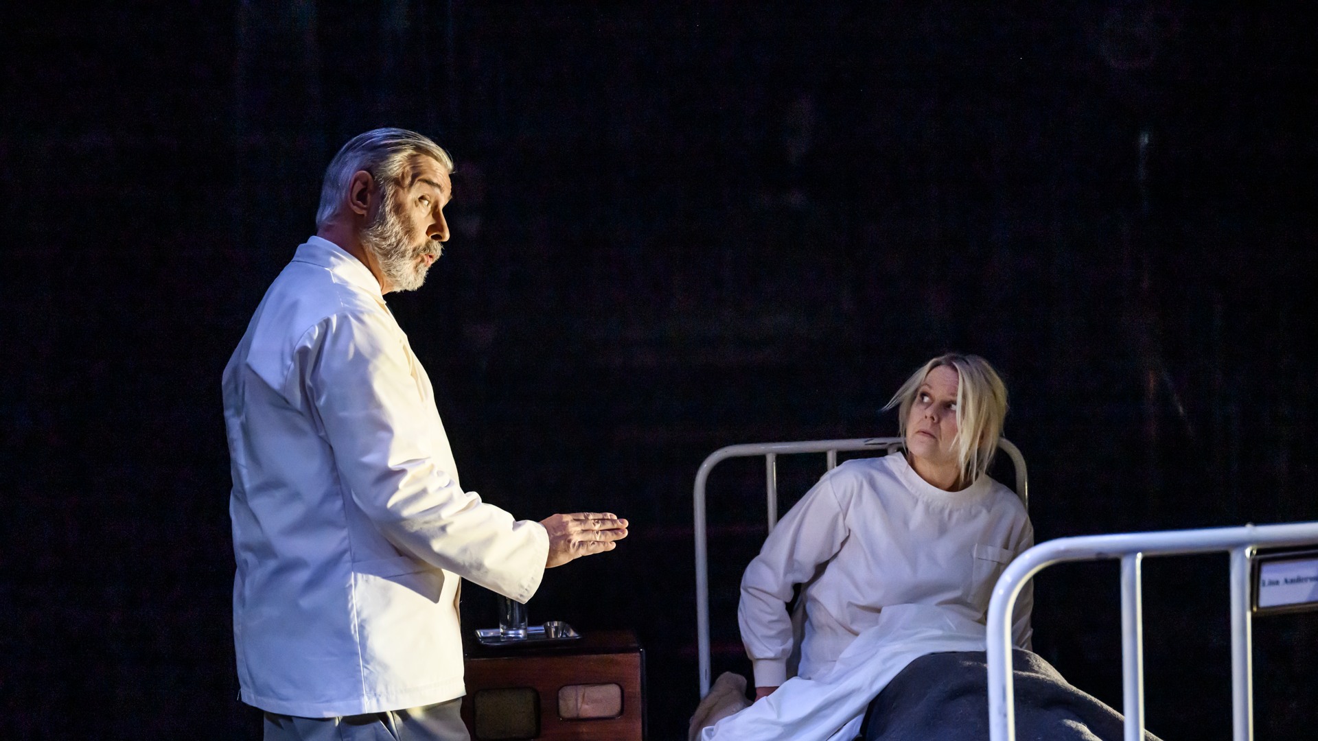 A woman is sitting in a hospital bed, a man in what looks like hospital clothes is talking to her.