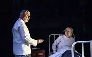 En kvinna sitter i en sjukhussäng, en man i vad som ser ut som sjukhuskläder talar med henne.