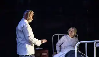 En kvinna sitter i en sjukhussäng, en man i vad som ser ut som sjukhuskläder talar med henne.