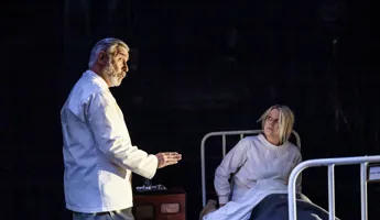 En kvinna sitter i en sjukhussäng, en man i vad som ser ut som sjukhuskläder talar med henne.