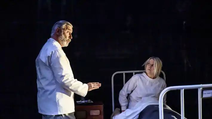 En kvinna sitter i en sjukhussäng, en man i vad som ser ut som sjukhuskläder talar med henne.