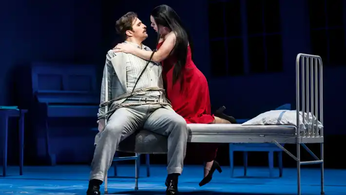 En man sitter fastbunden vid en säng, en kvinna i röd klänning håller om honom.