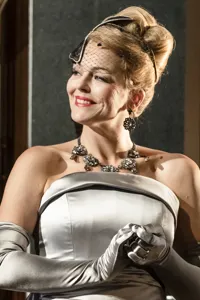 A smiling woman with updo hair, a small veil covering her face, a silver dress and long gloves.