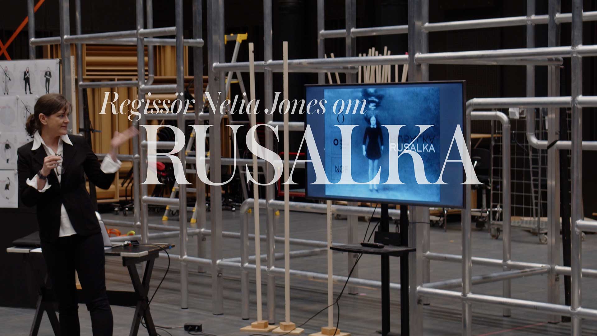 En kvinna står vid sidan av en TV-skärm, i bakgrunden syns byggnadsställningar.