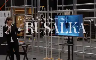 En kvinna står vid sidan av en TV-skärm, i bakgrunden syns byggnadsställningar.