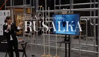 En kvinna står vid sidan av en TV-skärm, i bakgrunden syns byggnadsställningar.