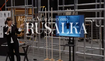 En kvinna står vid sidan av en TV-skärm, i bakgrunden syns byggnadsställningar.