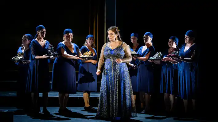 Several ladies dressed in blue on a stage, in the middle a woman with a gold-embellished blue dress.