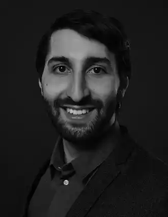 Närbild på en leende man med mörkt hår, skägg och mustasch; han bär en mörk skjorta och kavaj.