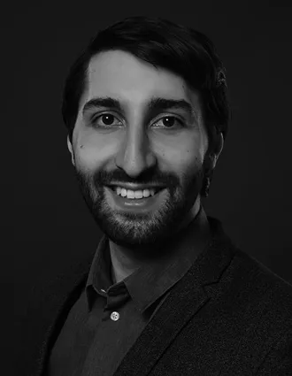 Närbild på en leende man med mörkt hår, skägg och mustasch; han bär en mörk skjorta och kavaj.