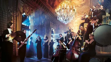 Several musicians with instruments in a grand room with a large chandelier, light shining in from the left.