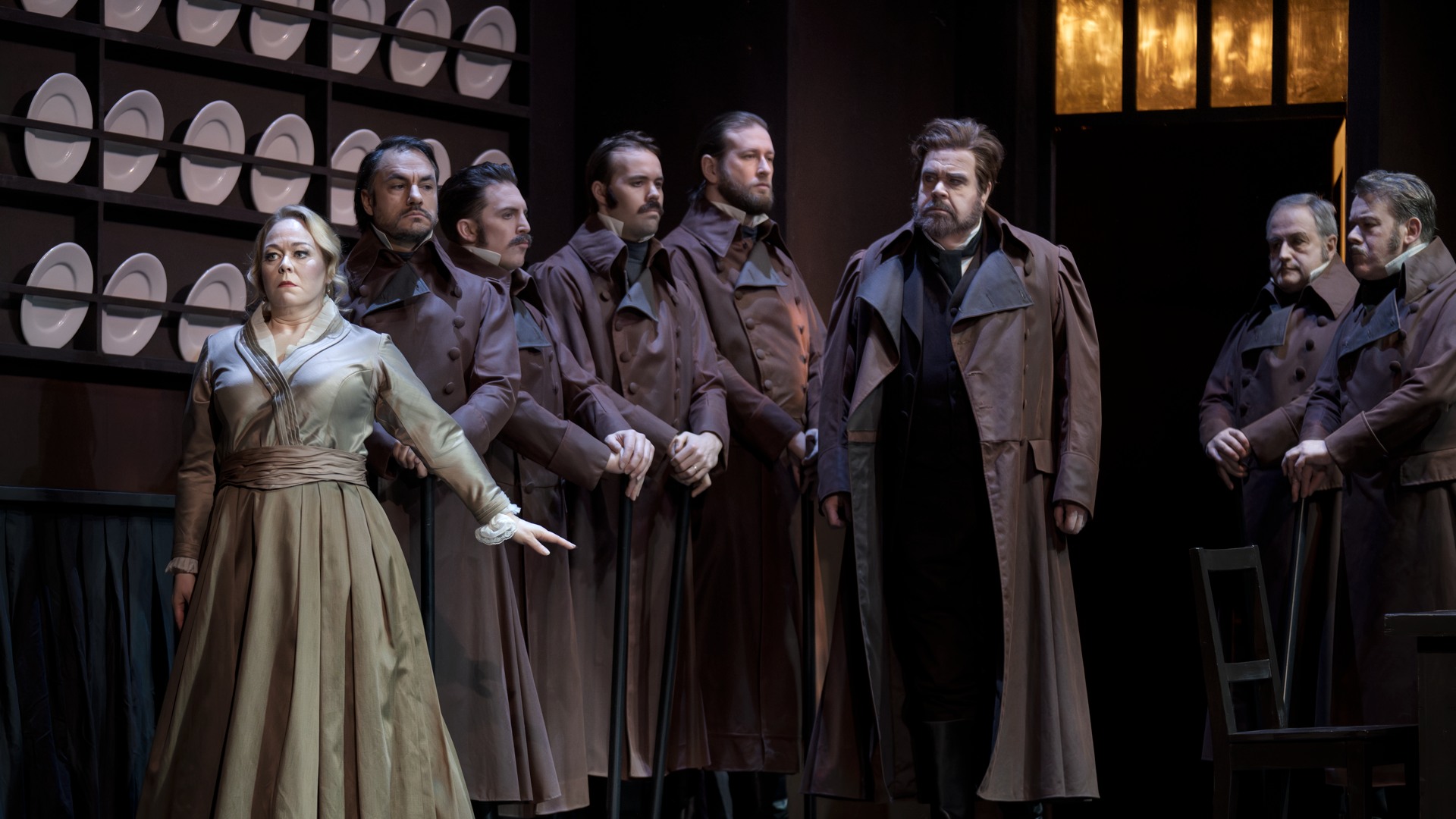 A group of people stand in a kitchen-like environment, plates lined up on the wall behind them.