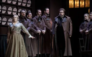 A group of people stand in a kitchen-like environment, plates lined up on the wall behind them.