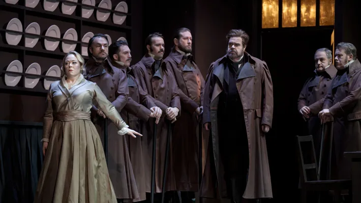 A group of people stand in a kitchen-like environment, plates lined up on the wall behind them.