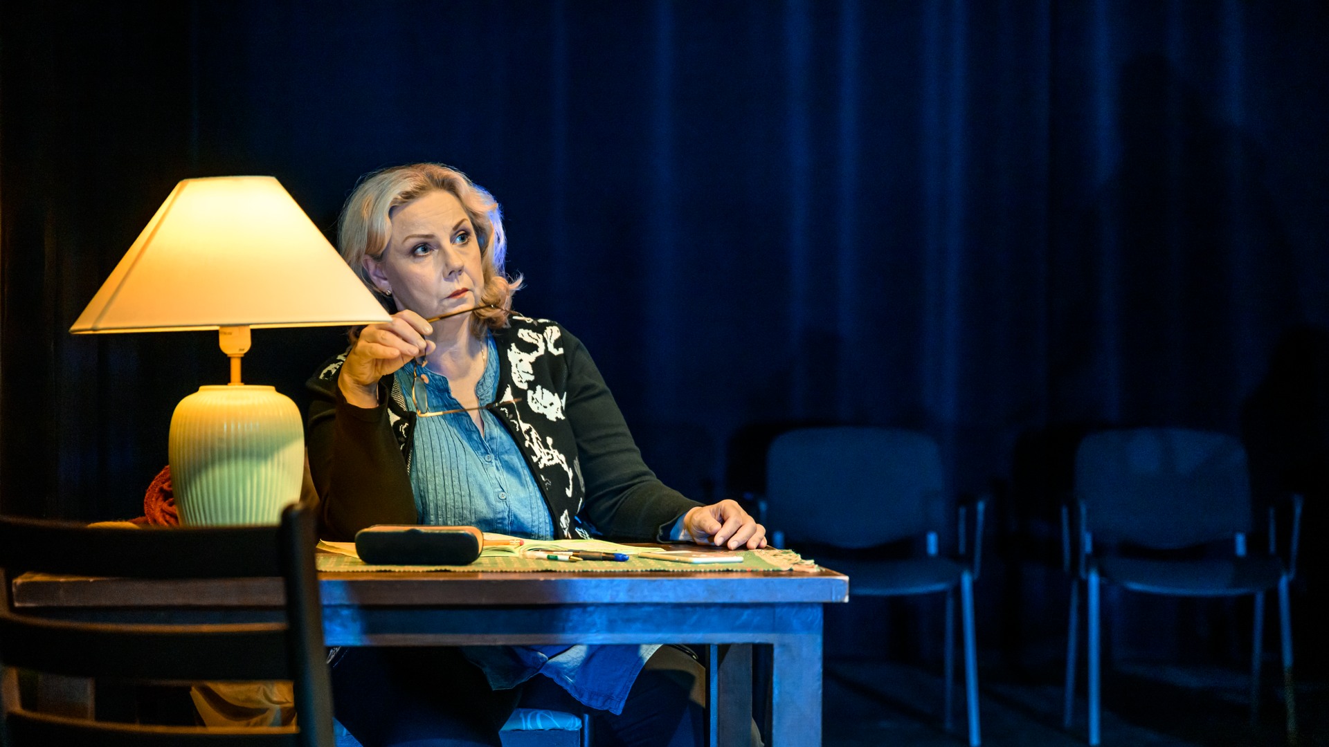 A woman, holding a pair of glasses, sits at a desk, on the table stands a lamp.