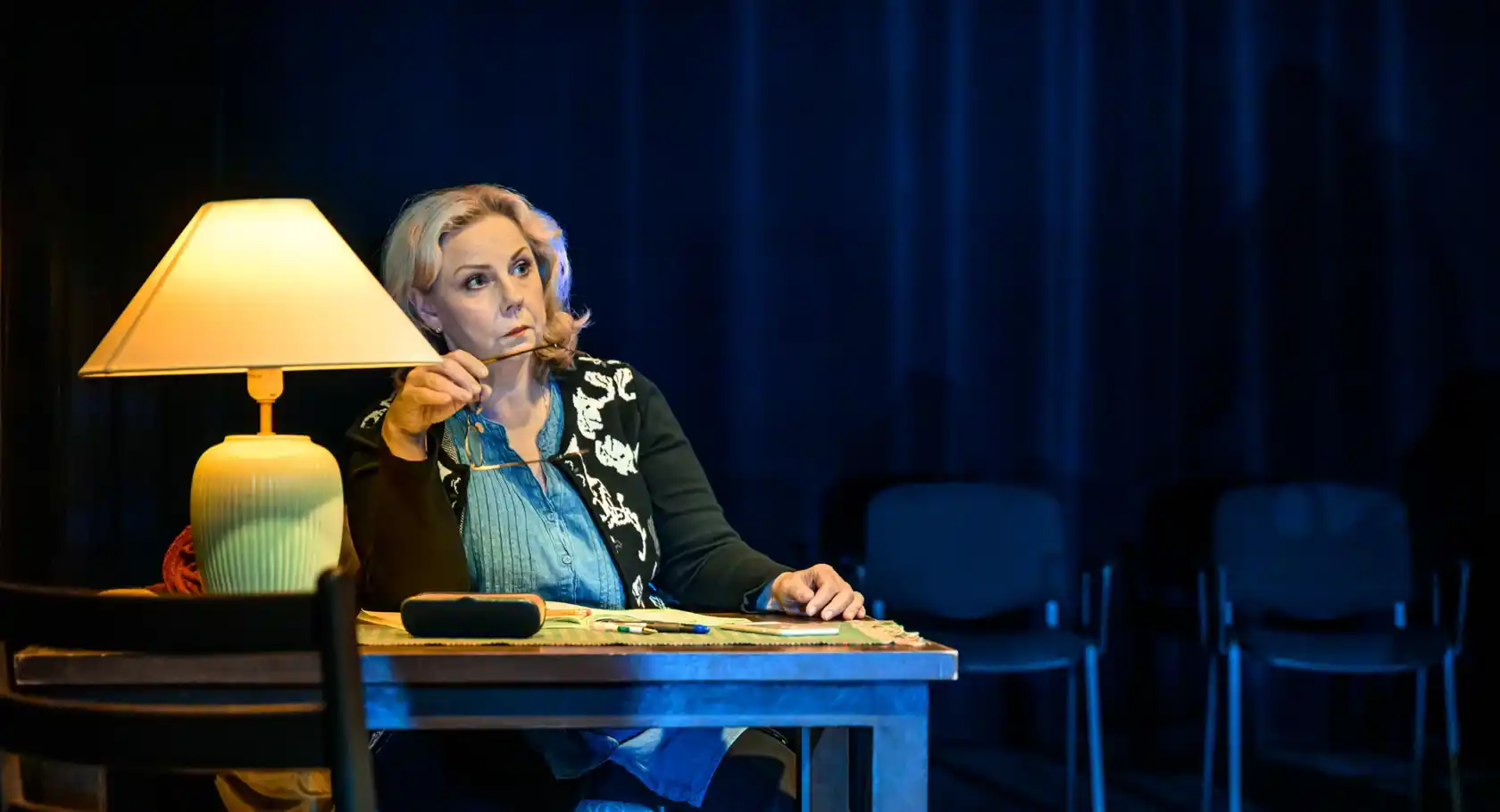 En kvinna, som håller i ett par glasögon, sitter vid ett skrivbord, på bordet står en lampa.