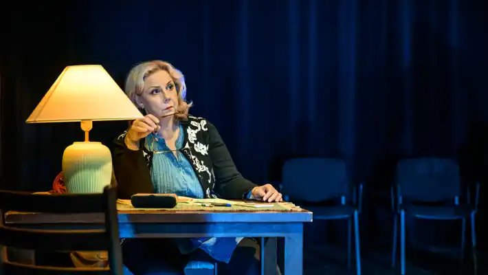 En kvinna, som håller i ett par glasögon, sitter vid ett skrivbord, på bordet står en lampa.