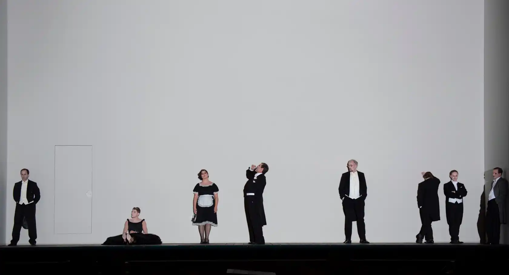 Against a large, blank white wall stands a group of people dressed in white and black.