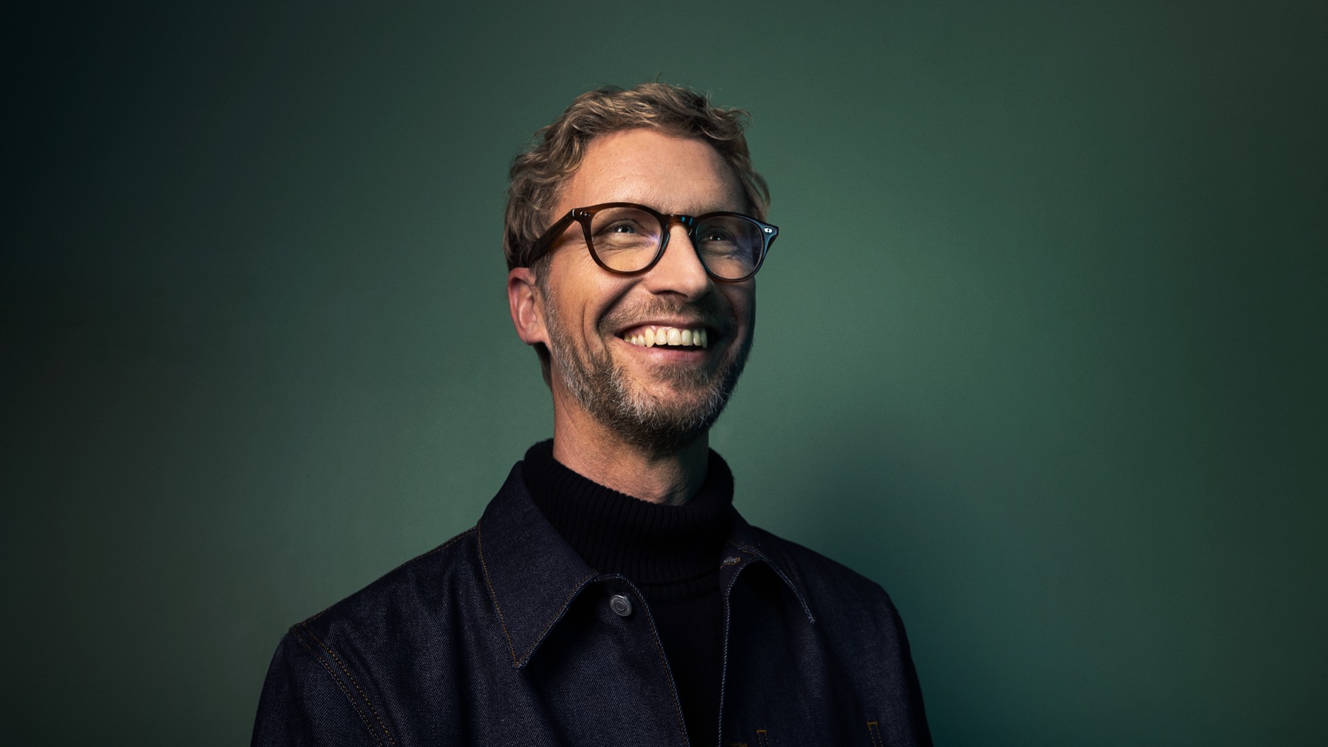 A laughing man in glasses in front of a dark green background.