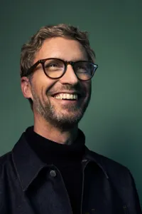 En skrattande man i glasögon framför en mörkt grön bakgrund.