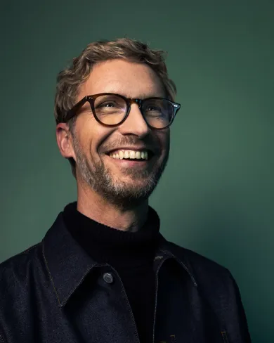 A laughing man in glasses in front of a dark green background.