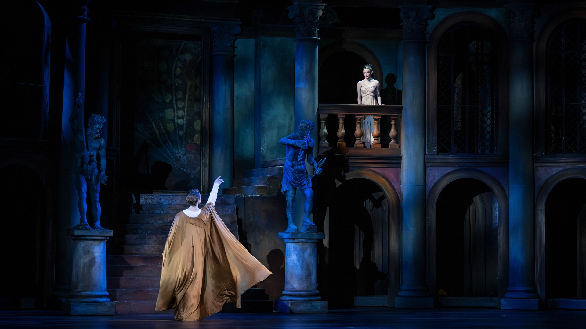 En man med utsträckt arm står nedanför en balkong, på balkongen står en ung kvinna.