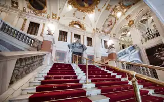 A large, wide staircase with red carpet leads up into an ornate stairwell.