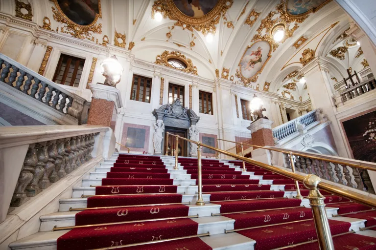 A large, wide staircase with red carpet leads up into an ornate stairwell.