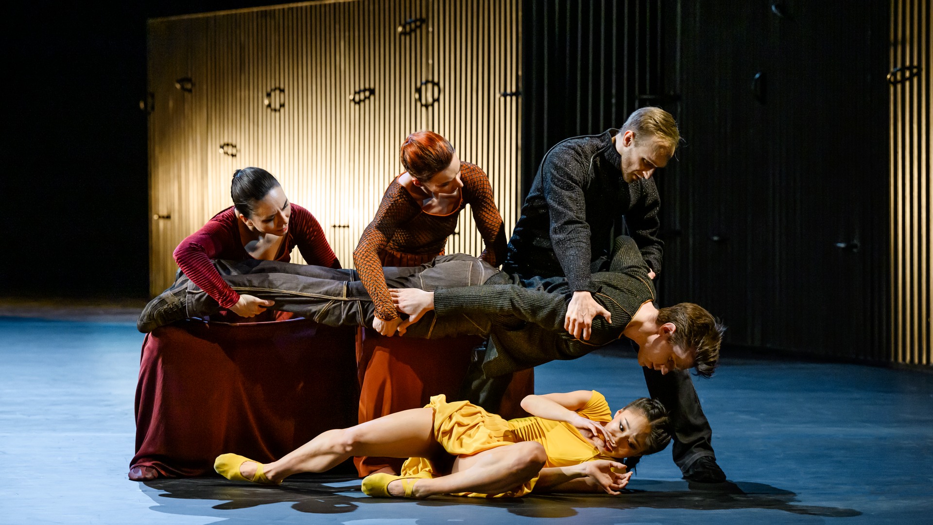 Två kvinnor och en man håller en man svävande över en rädd kvinna som ligger på golvet.